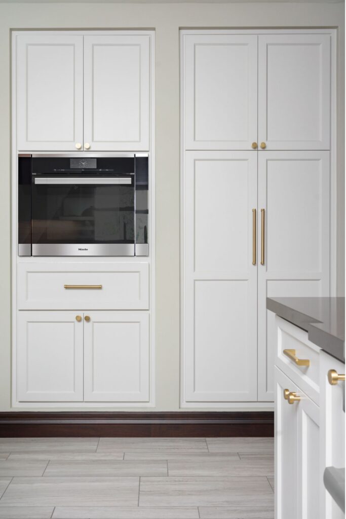 Built-in wall oven with floor-to-ceiling white shaker cabinetry, brass pulls and light tile flooring