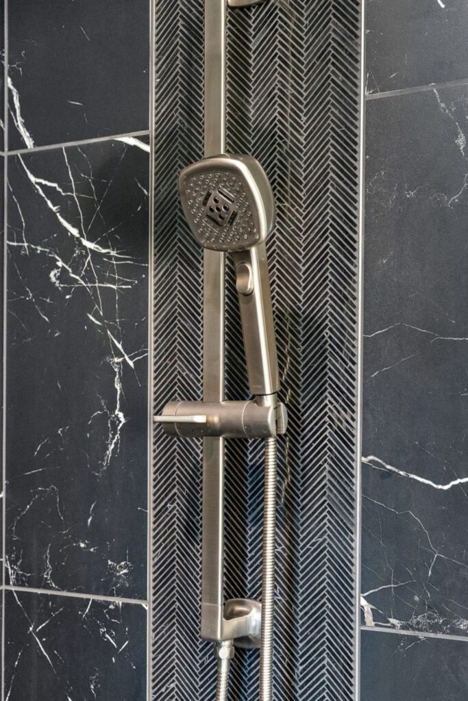 Handheld shower fixture mounted on black herringbone accent tile between black veined wall tiles