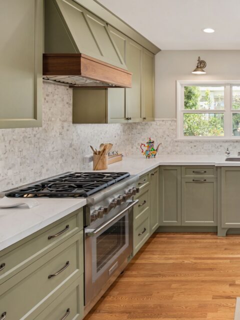 Sage green kitchen remodel with stainless range, wood-trimmed hood, mosaic backsplash, white counters and hardwood flooring.