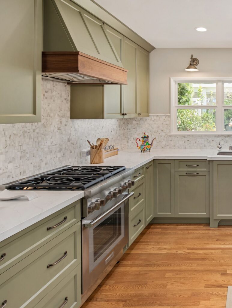 Sage green kitchen remodel with stainless range, wood-trimmed hood, mosaic backsplash, white counters and hardwood flooring.