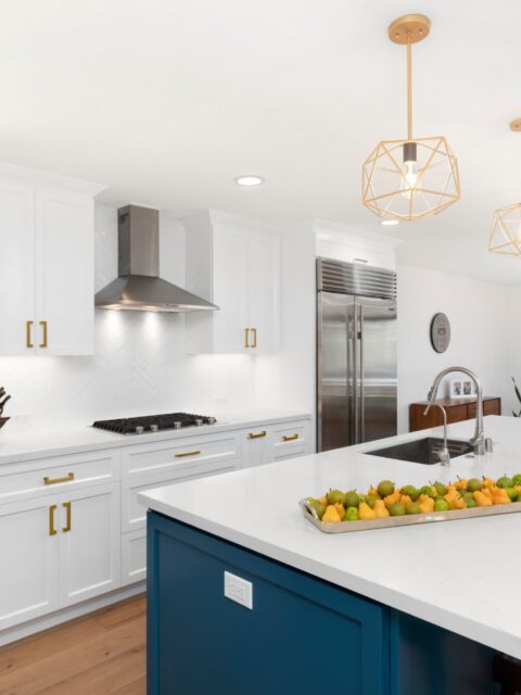 Oceanside whole-house remodel kitchen with blue island, white cabinetry, brass pendants, herringbone backsplash, and stainless appliances.
