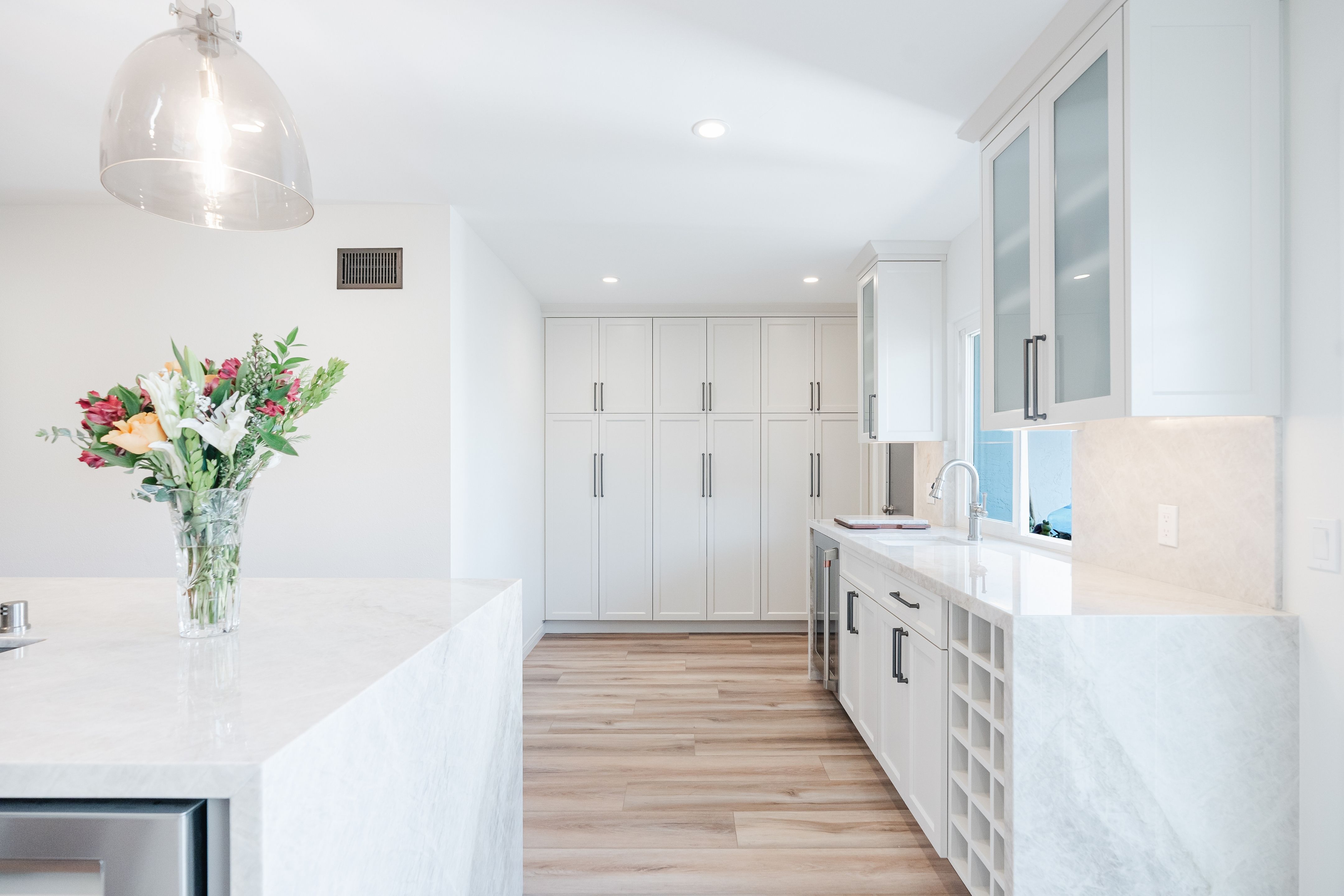 Kitchen with white shaker cabinetry, waterfall stone counters, glass-front uppers, pantry storage and wood-look flooring.