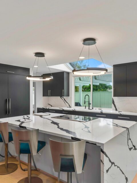 Modern kitchen with dark cabinetry, waterfall island, induction cooktop, slab backsplash and ring pendant lights.