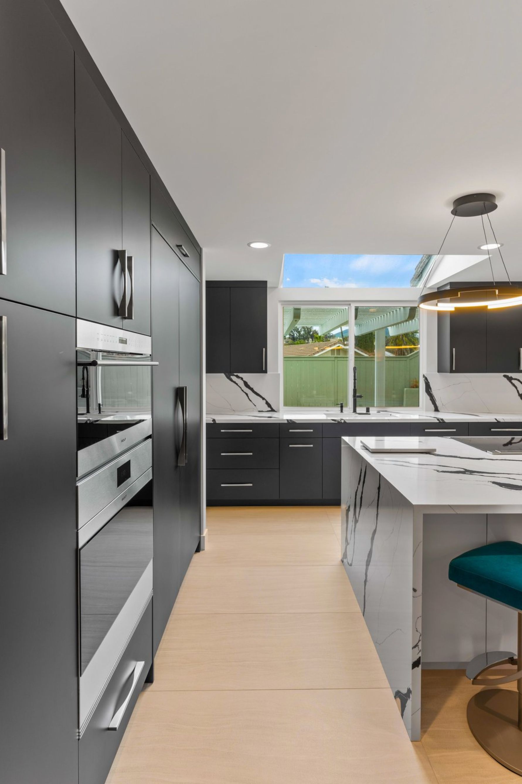 Modern kitchen with dark cabinetry, waterfall island, built-in ovens, skylight and ring pendant lighting.