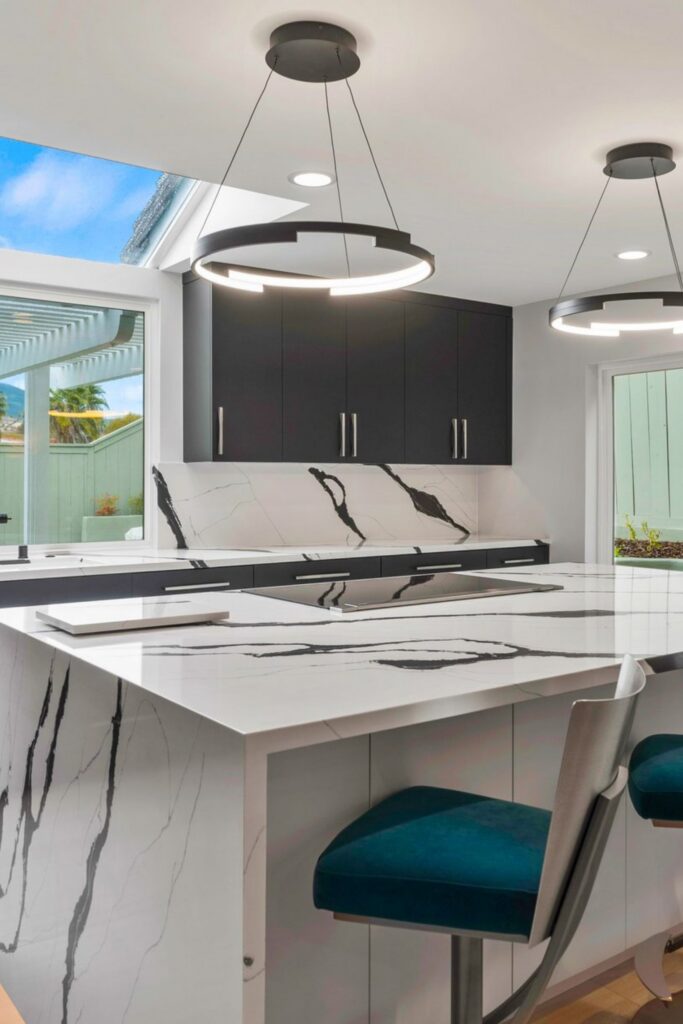 Waterfall kitchen island with veined countertop, teal stools, dark cabinetry and circular pendant lighting.