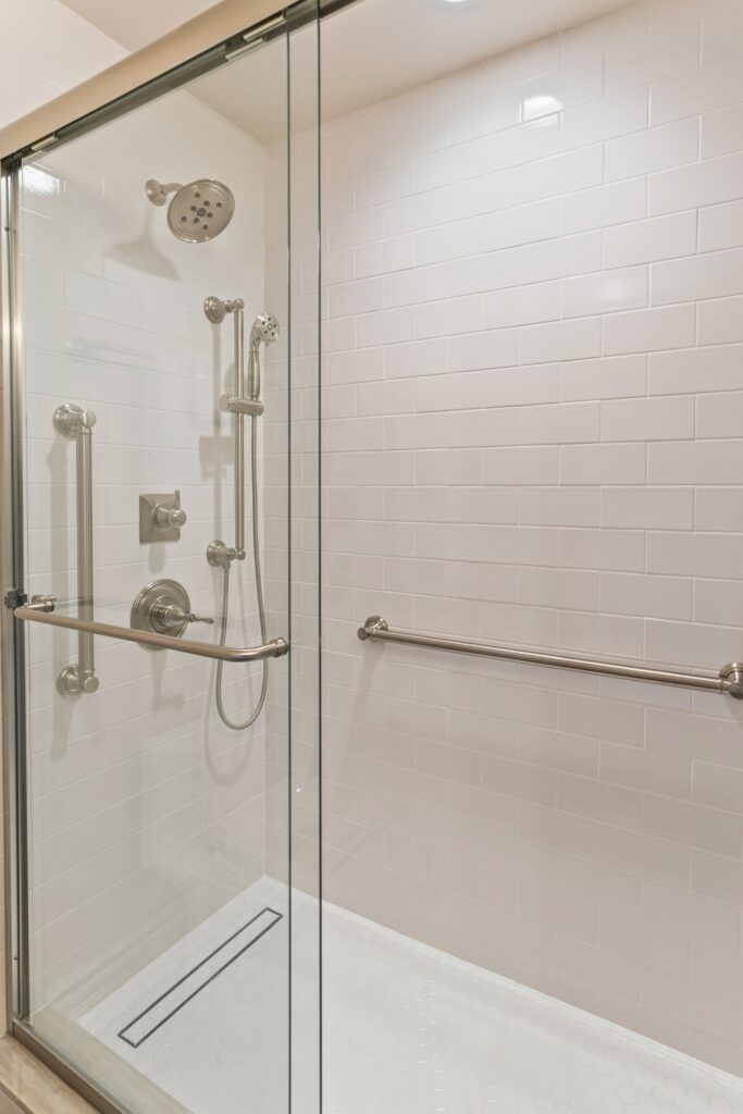 Walk-in shower with white subway tile, glass enclosure, handheld showerhead and grab bars.