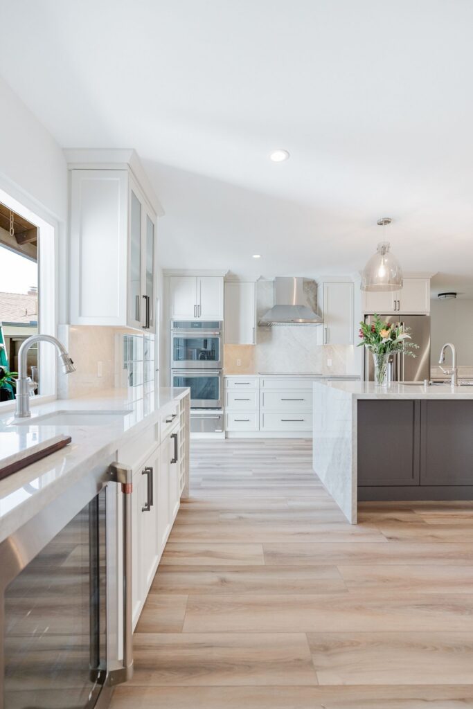 Kitchen with dark island, white shaker cabinets, stainless hood and double ovens, pendant lights and wood-look flooring.
