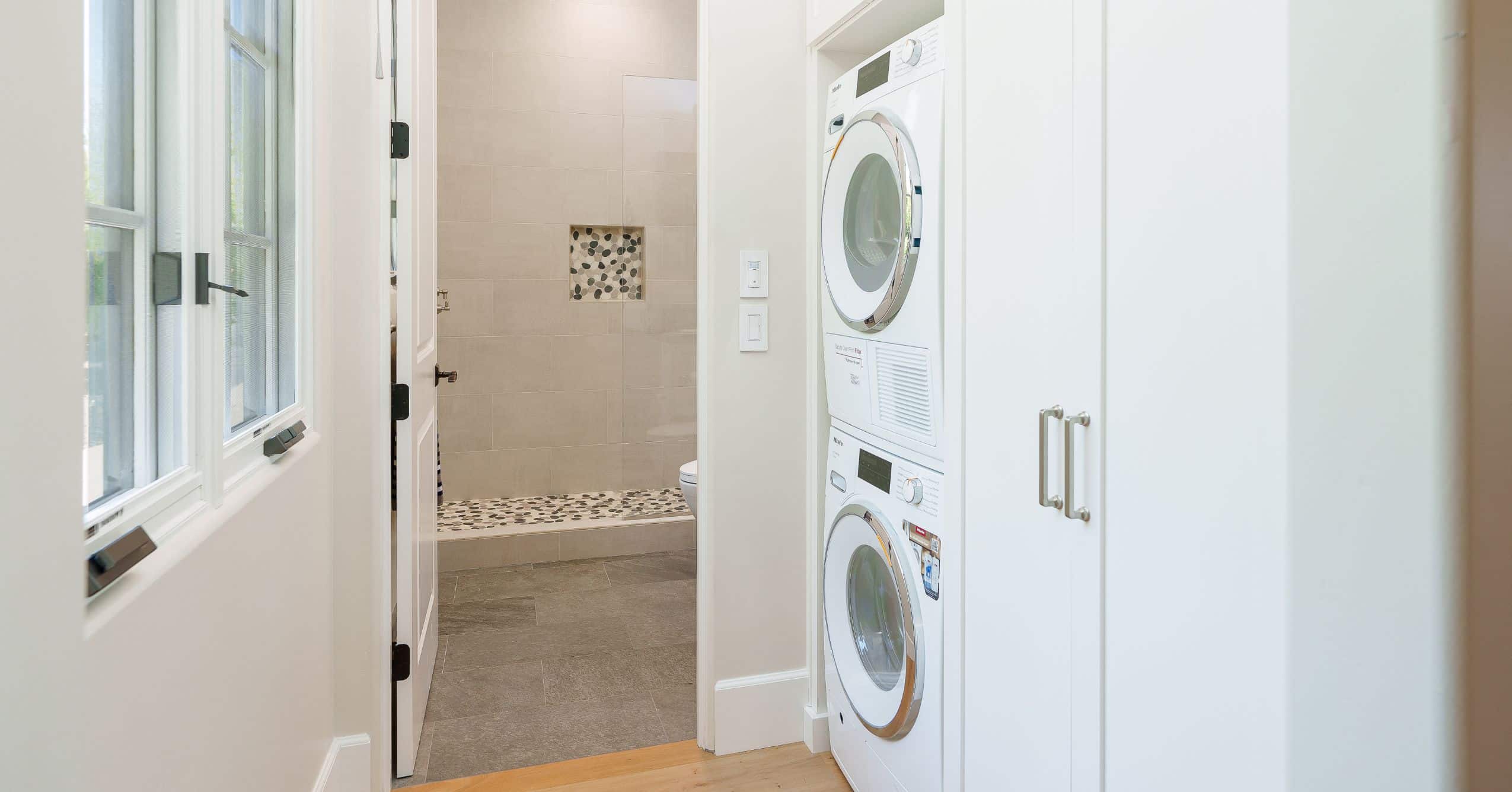 Hallway with stacked washer and dryer, white cabinetry, and entrance to a full tiled bathroom.