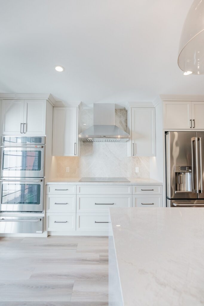 Cooking wall with white shaker cabinetry, stainless range hood, double ovens, refrigerator and slab backsplash.