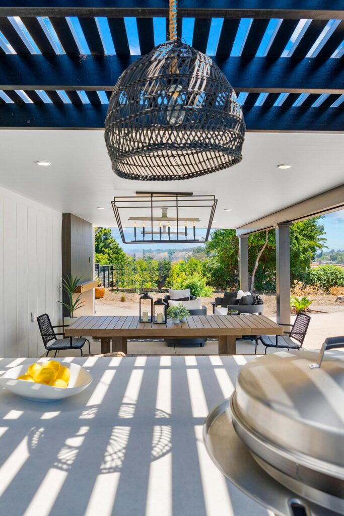 Covered dining area viewed from outdoor kitchen with wood table, pendant lighting and lounge seating beyond