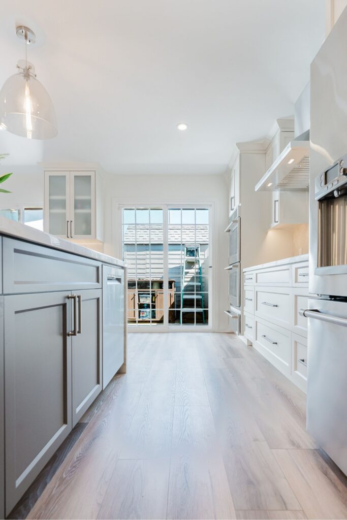 Kitchen aisle with dark island, white cabinetry, stainless appliances, glass patio door and light wood-look flooring.