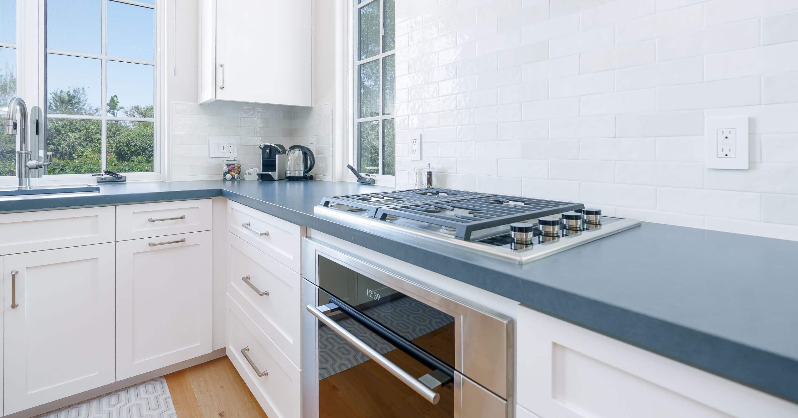 Close-up of ADU kitchen with white cabinets, dark countertops, gas cooktop, wall oven, and white tile backsplash.
