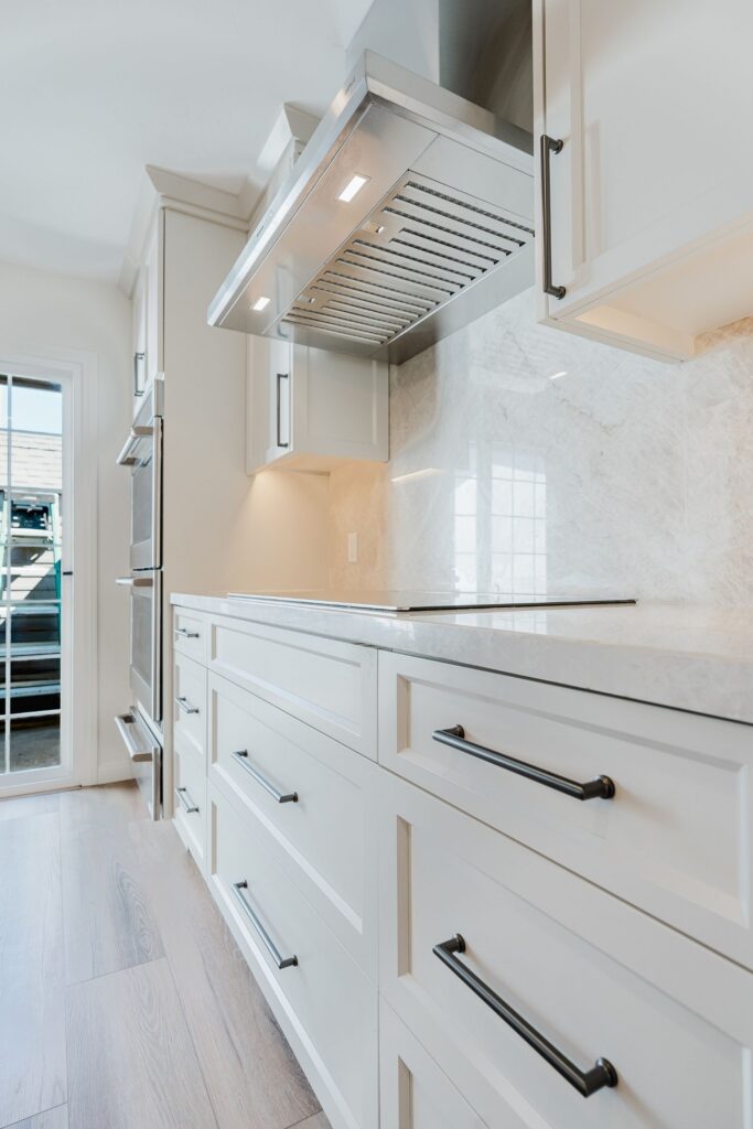 Stainless range hood above slab backsplash and cooktop with white shaker drawers and dark cabinet pulls.