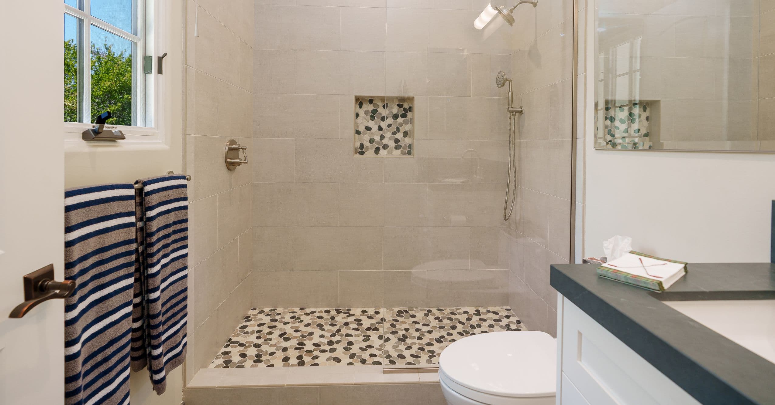 Full ADU bathroom with glass-enclosed walk-in shower, pebble tile floor, white vanity, and dark countertop.