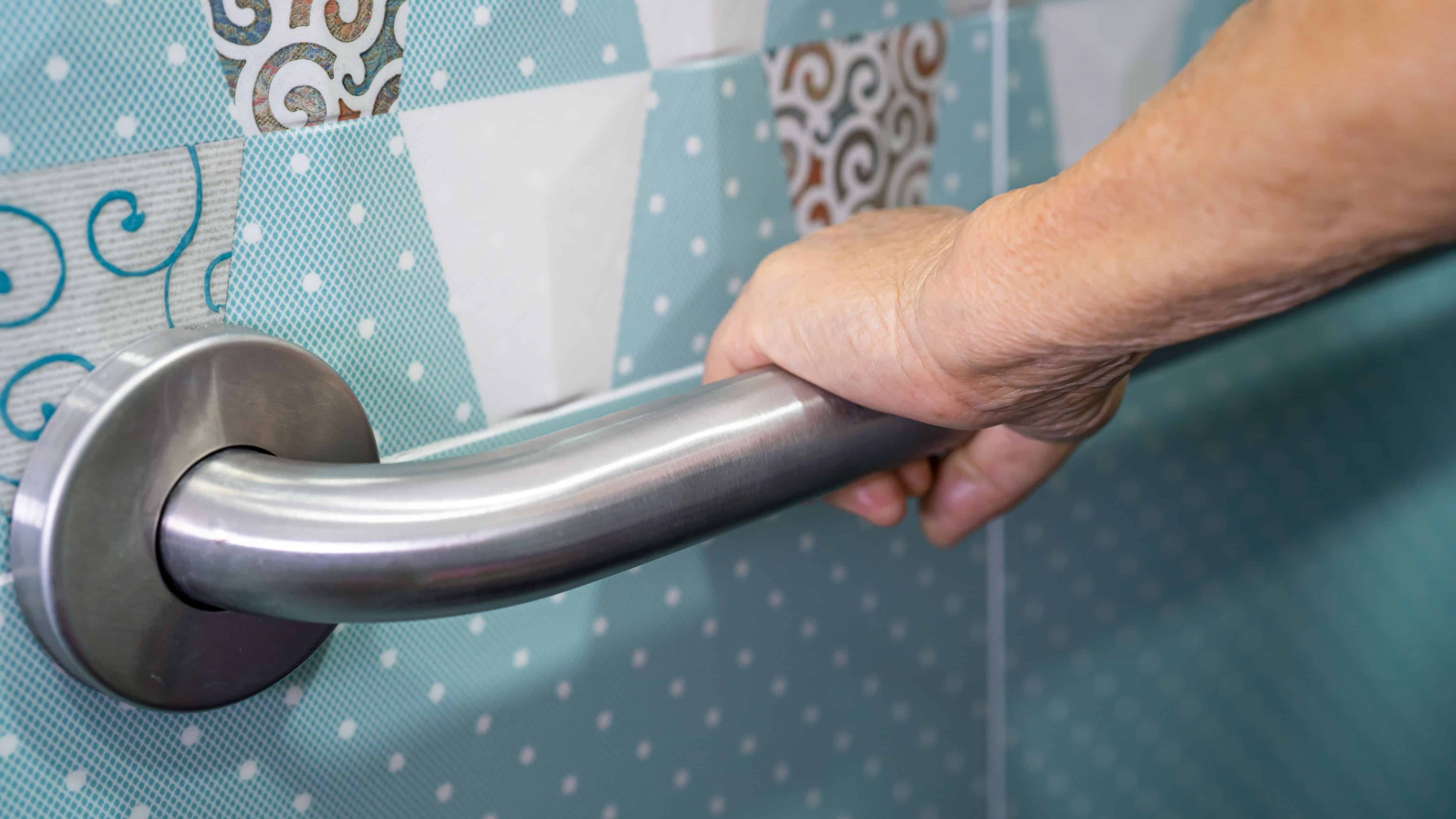 Older adult holding a stainless-steel bathroom grab bar installed beside a shower for added stability and support.