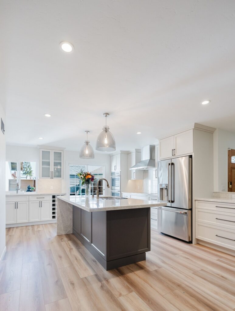 Open kitchen remodel with dark island, white shaker cabinetry, stone counters, stainless appliances and pendant lights.