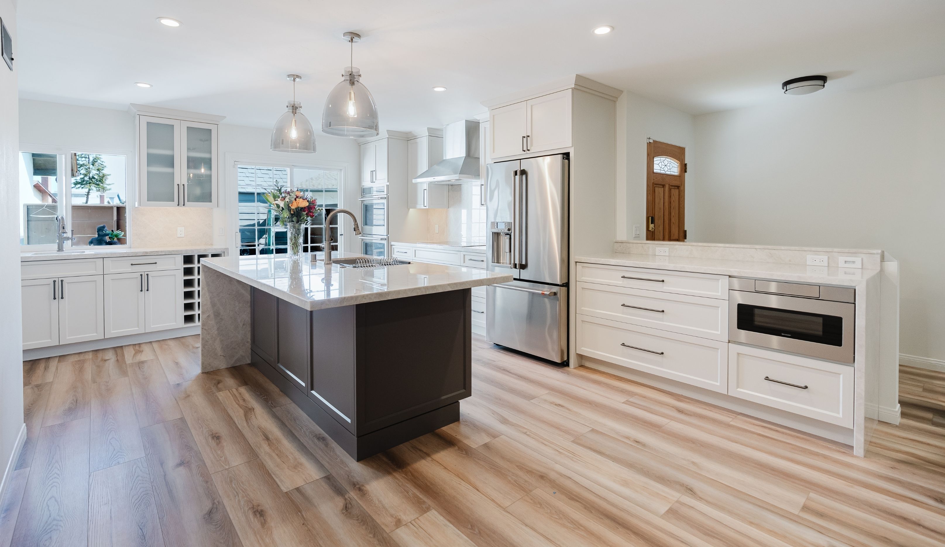 Wide kitchen remodel with waterfall island, white cabinetry, stainless appliances, built-in storage and wood-look flooring.