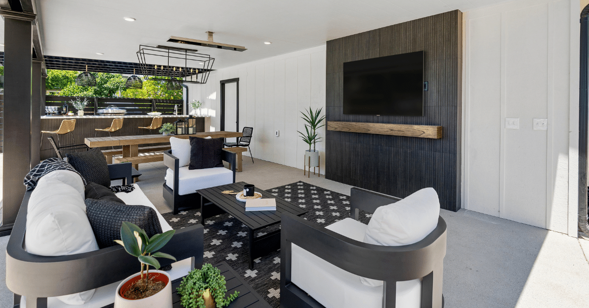Covered outdoor living room with dark sofas and white cushions, a patterned rug and a TV on a dark slatted wall, with a dining table and outdoor kitchen bar behind.