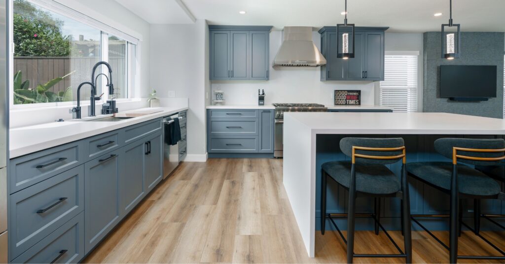Remodeled kitchen with blue-gray cabinetry, white quartz countertops, large island, pendant lights, and wood-look flooring.