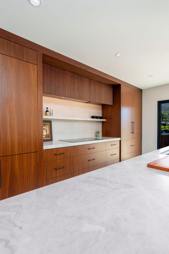 Walnut cabinet wall with induction cooktop, fluted wood hood panel, stone backsplash and floating shelf behind quartzite island