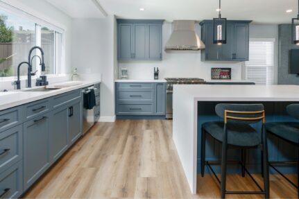 Remodeled kitchen with blue-gray cabinetry, white quartz countertops, large island, pendant lights, and wood-look flooring.