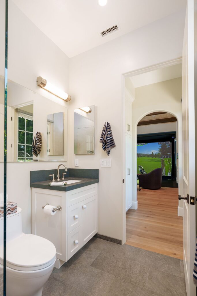 Guest bathroom with a white shaker vanity, charcoal countertop, golf-ball knobs and gray tile, opening to the simulator room