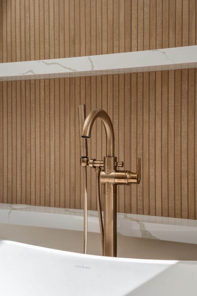Gold-tone freestanding tub filler with handheld sprayer against a vertical wood-slat panel and veined quartz shelves