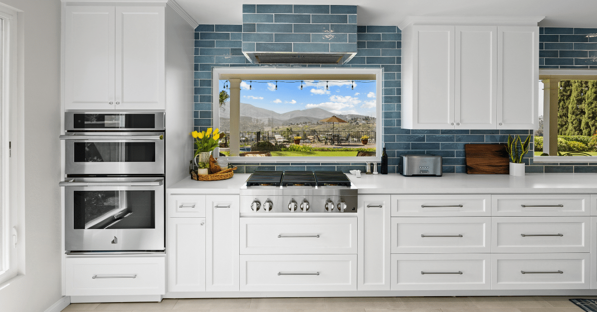 White kitchen with blue subway tile backsplash, stainless steel appliances, and a cooktop beneath a large window.