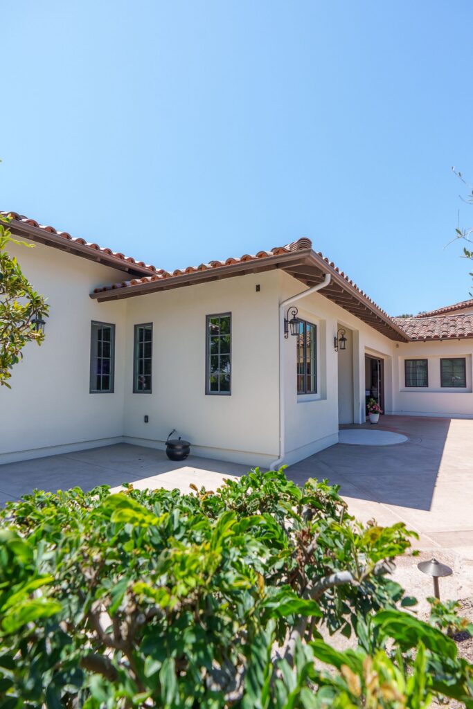 Side view of a stucco ADU with tall divided-lite windows, wall lanterns, a clay tile roof and a concrete patio