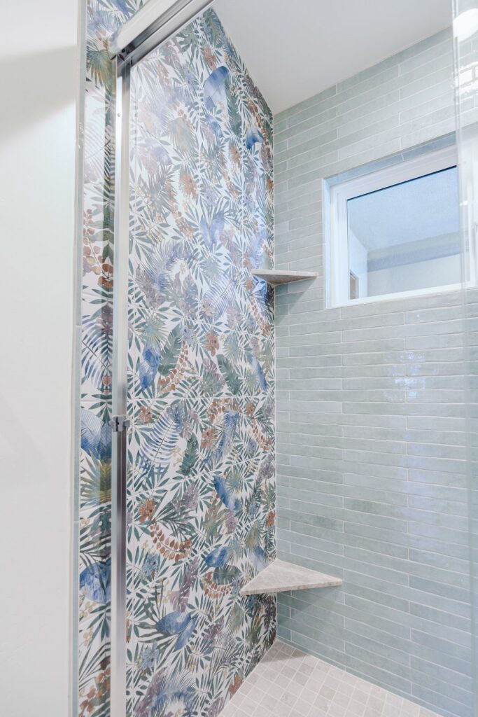 Shower with a botanical-print tile accent wall, green subway tile, stone corner shelves and a mosaic tile floor