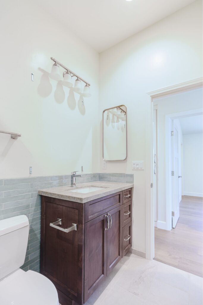 Dark wood Shaker vanity with a stone top, undermount sink, green tile wainscot, four-light fixture and rounded mirror