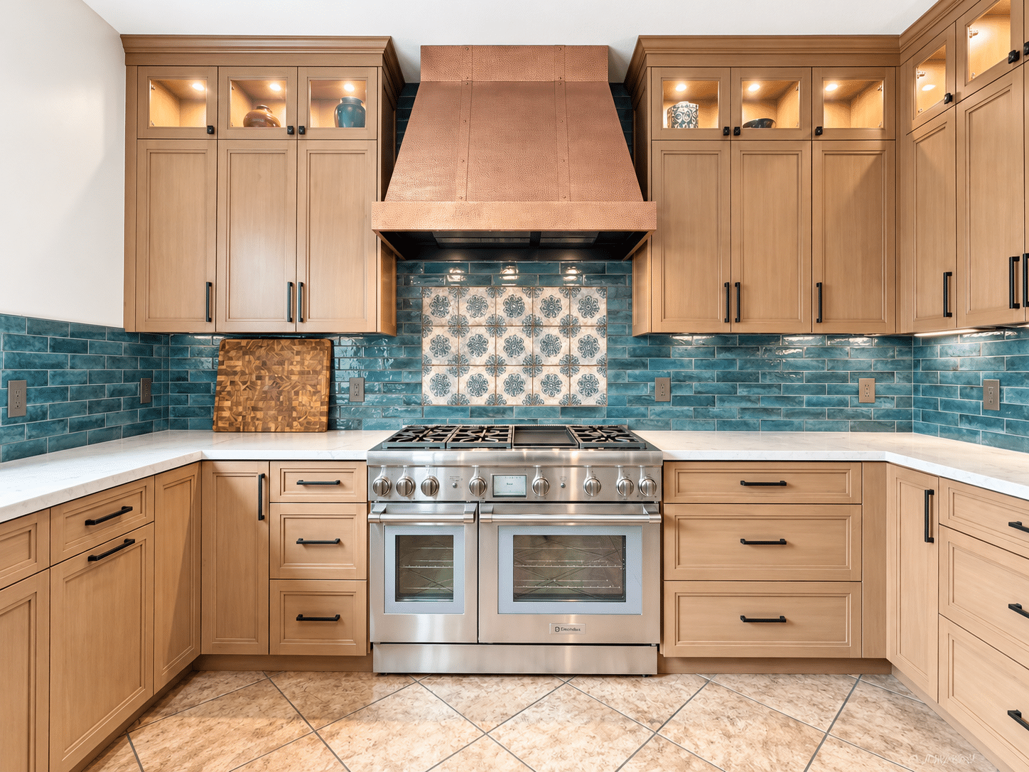 Straight-on view of a stainless steel range with six burners and a griddle beneath a copper hood, backed by a blue-and-cream patterned tile panel set into glossy teal subway-style tile, between light wood cabinets with dark bronze pulls.