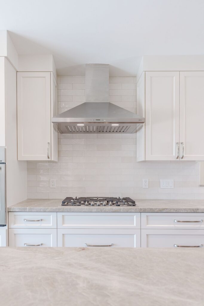Gas cooktop under a stainless chimney hood, with white glazed tile backsplash and white Shaker drawers below