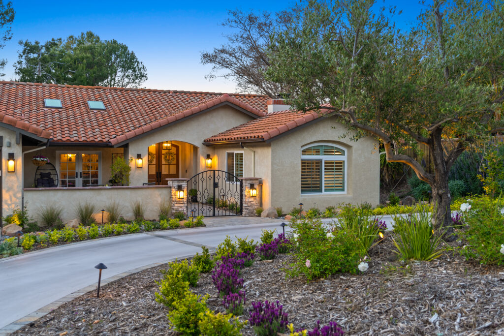 Spanish-style home at dusk with a walled front courtyard, a wrought iron gate between stone pillars, lantern sconces, a curved driveway and mulched beds of lavender beneath an olive tree.