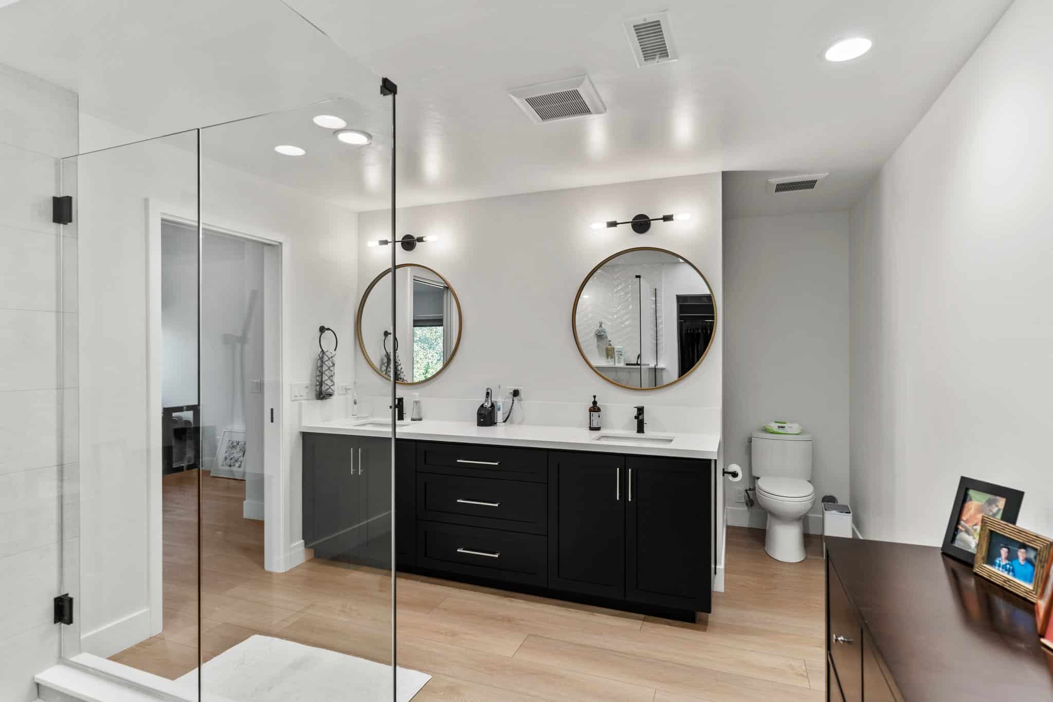 Contemporary bathroom with black double-sink vanity, round mirrors, glass shower enclosure, light wood flooring, and separate toilet area.