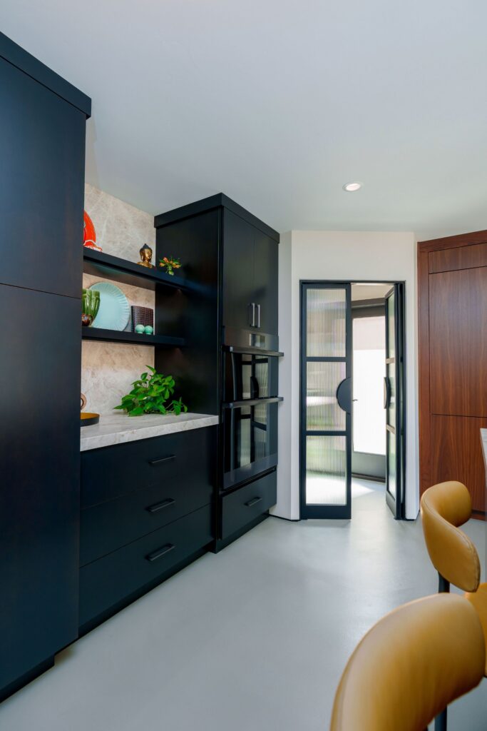 Black kitchen cabinetry with stacked wall ovens, open shelving, stone backsplash and reeded-glass steel-framed doors