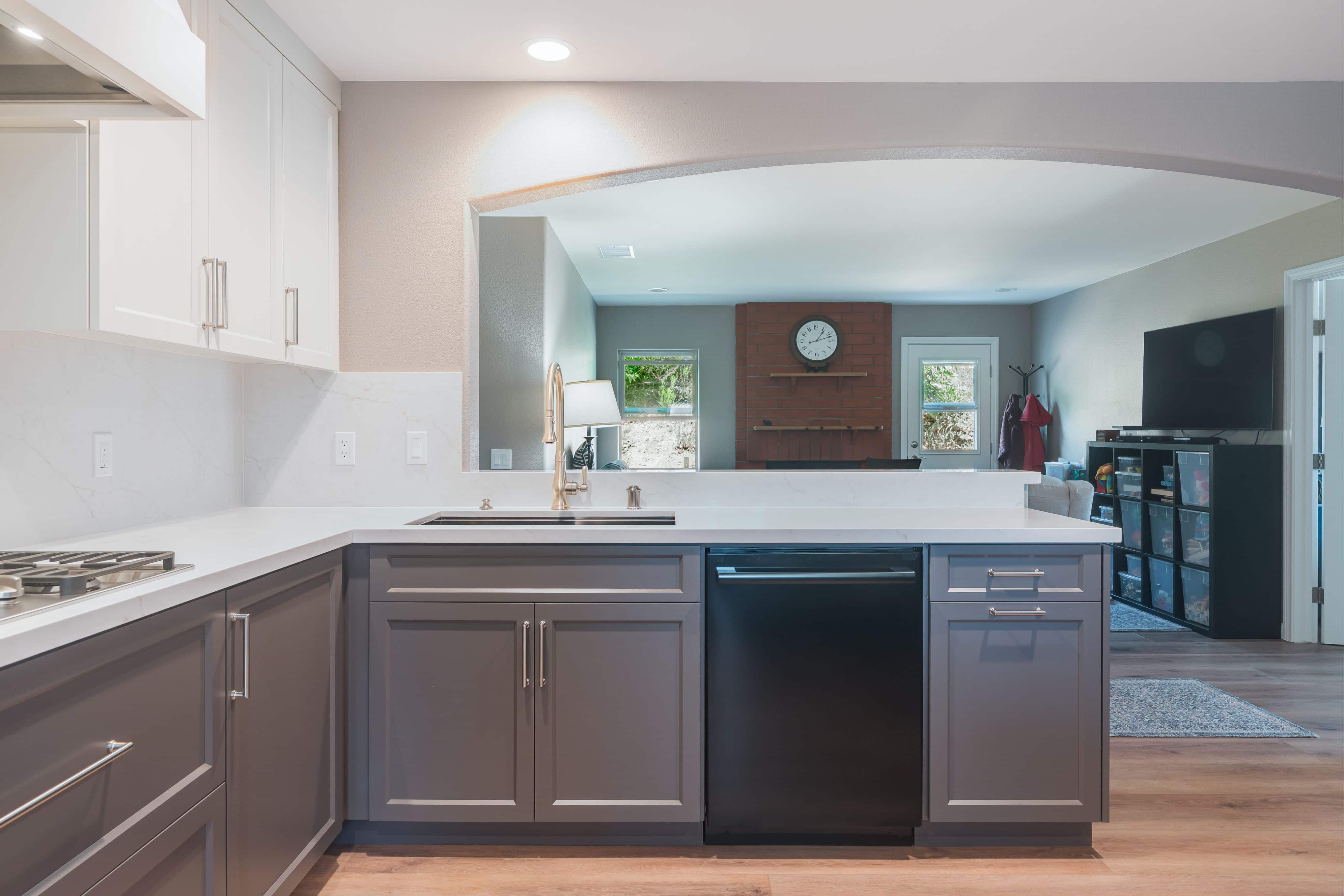 Kitchen sink run with gray shaker cabinets, white quartz counters, black dishwasher and arched opening to the family room