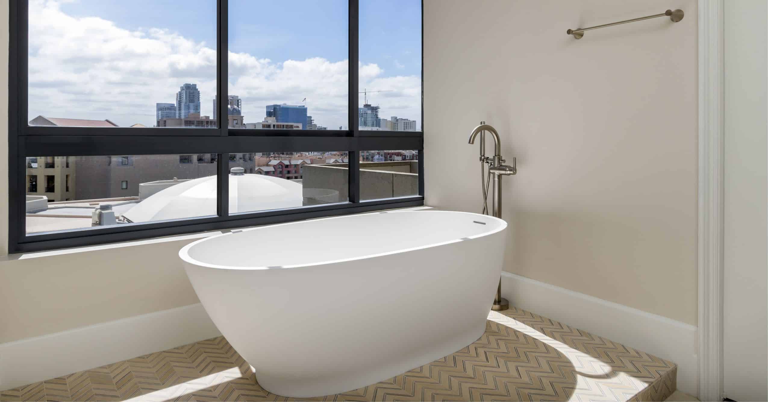 Freestanding white bathtub beside a large black-framed window with a city view and patterned tile flooring.