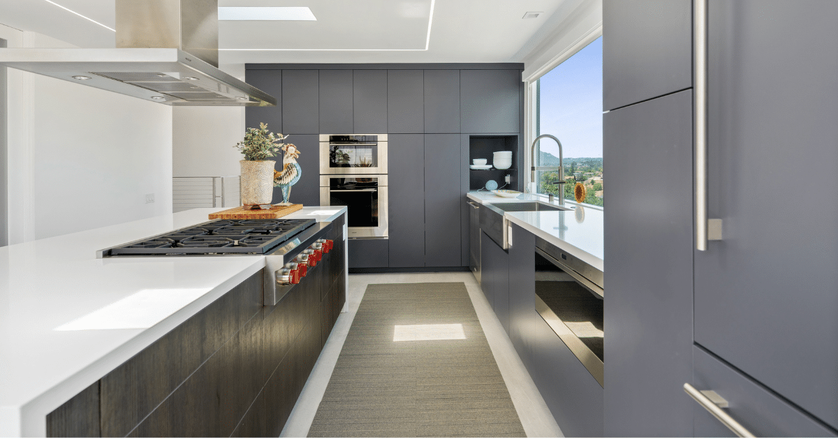 Modern gray kitchen with flat-panel cabinetry, long island cooktop, white countertops, and large window views.