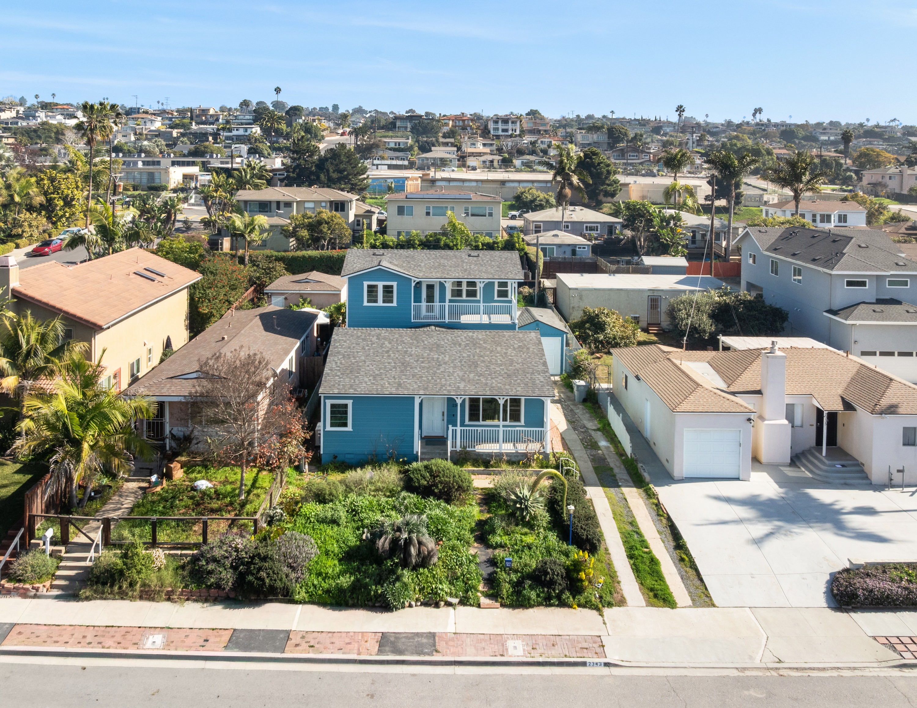 Aerial view of a blue house with a white-trimmed front porch, a single-story front section and a two-story rear section with an upper balcony, on a narrow lot among neighboring homes.