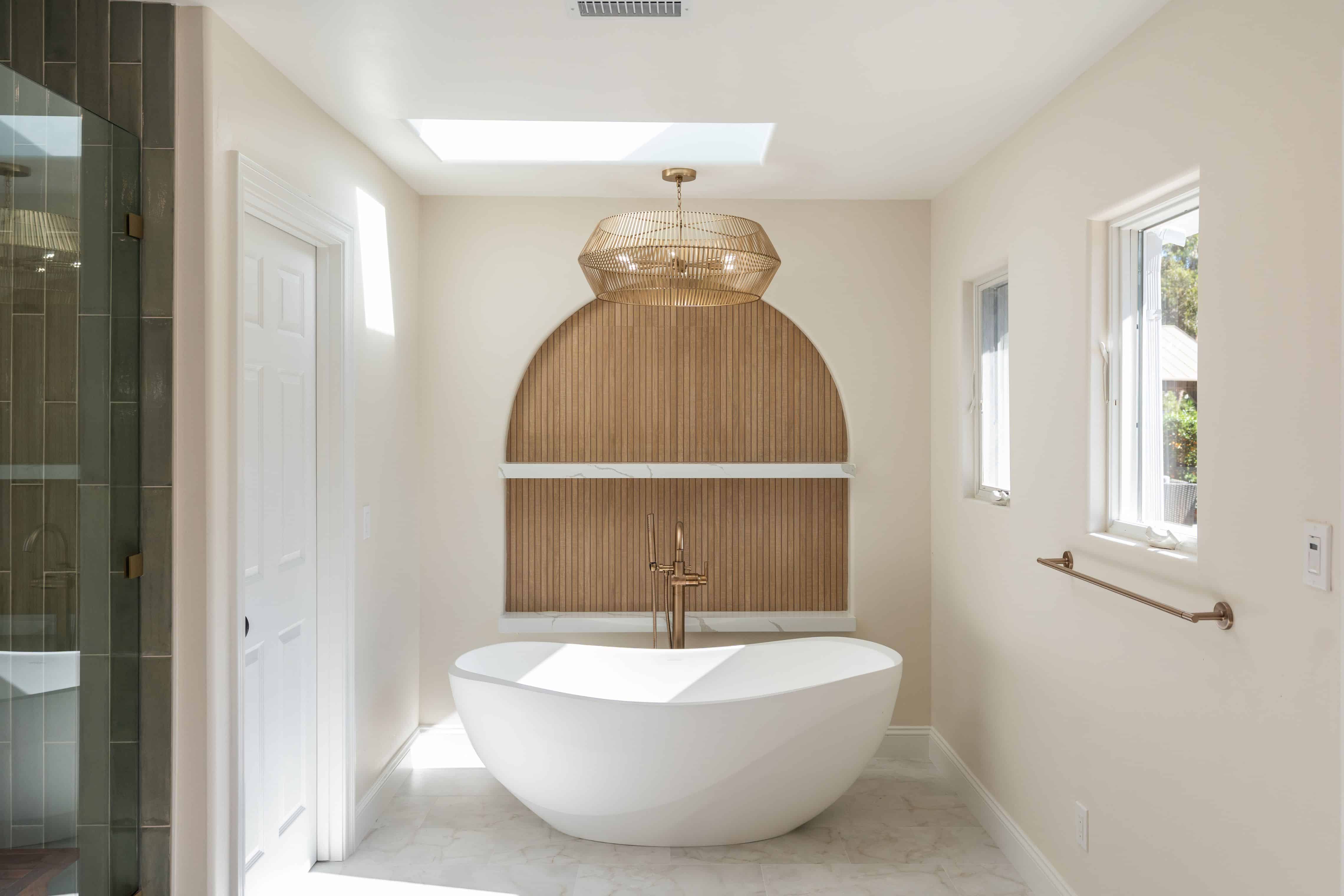 Freestanding soaking tub in arched wood-slat niche with quartz ledges, gold cage pendant, skylight and side windows