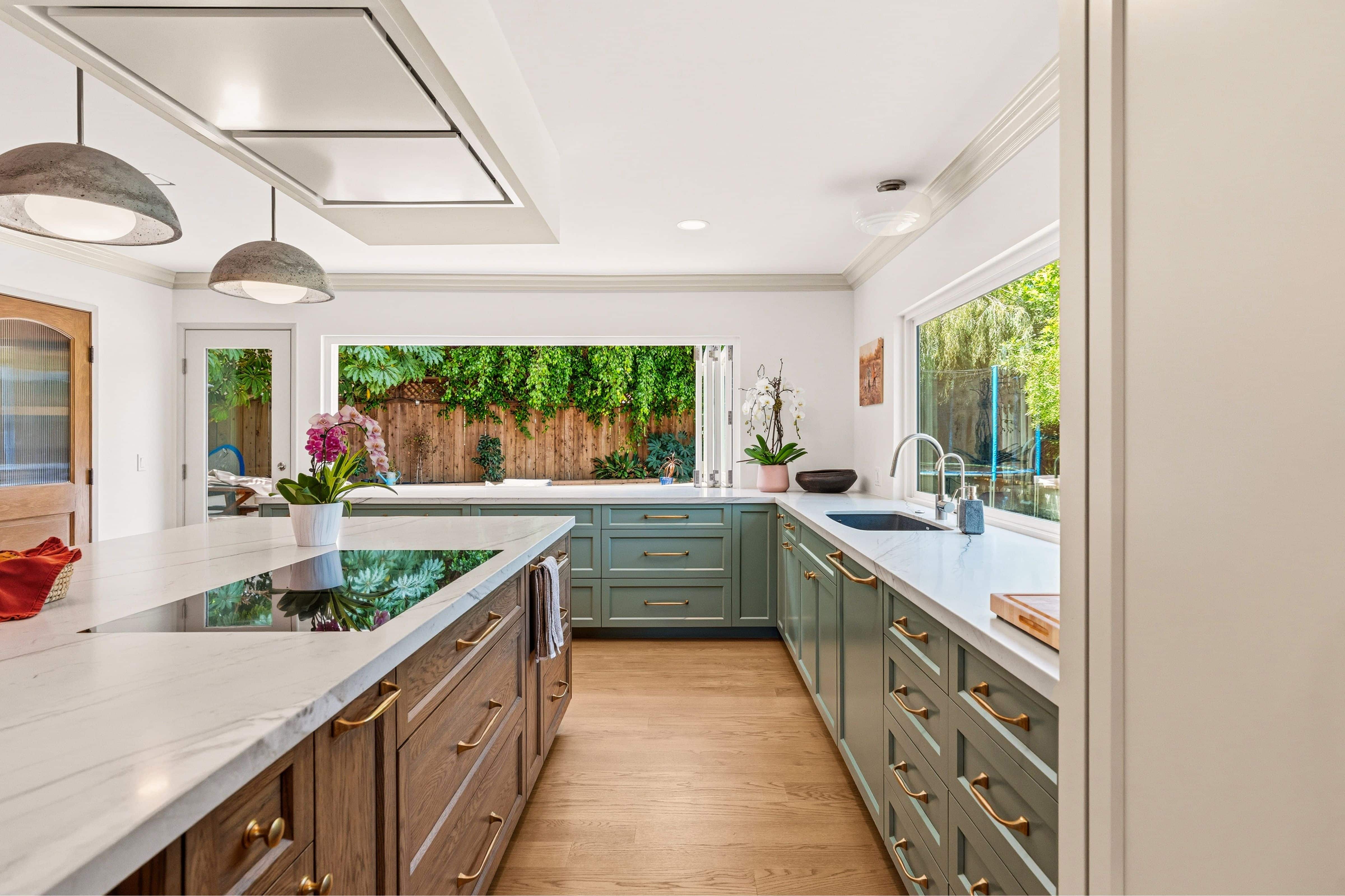 Galley view of green shaker cabinets, wood island with flush cooktop, ceiling-mounted hood and bi-fold window to side yard