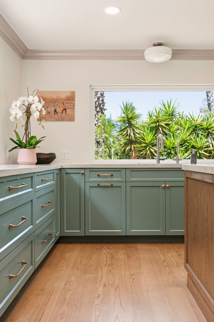 Corner of green shaker base cabinets with brass pulls, wood-plank flooring and a sink window framing yucca plants