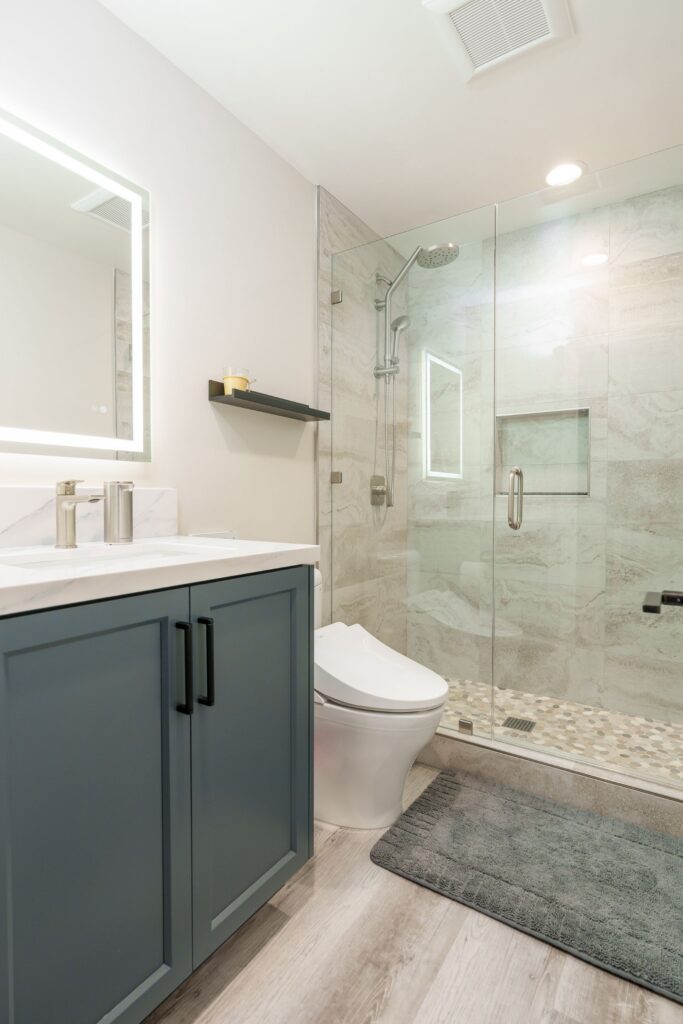 Bathroom with blue shaker vanity, black pulls, LED backlit mirror, frameless glass shower, stone-look tile and pebble floor