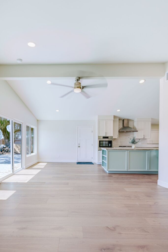 Open living area with a vaulted ceiling, exposed beam, ceiling fan and plank flooring leading to a sage green kitchen island