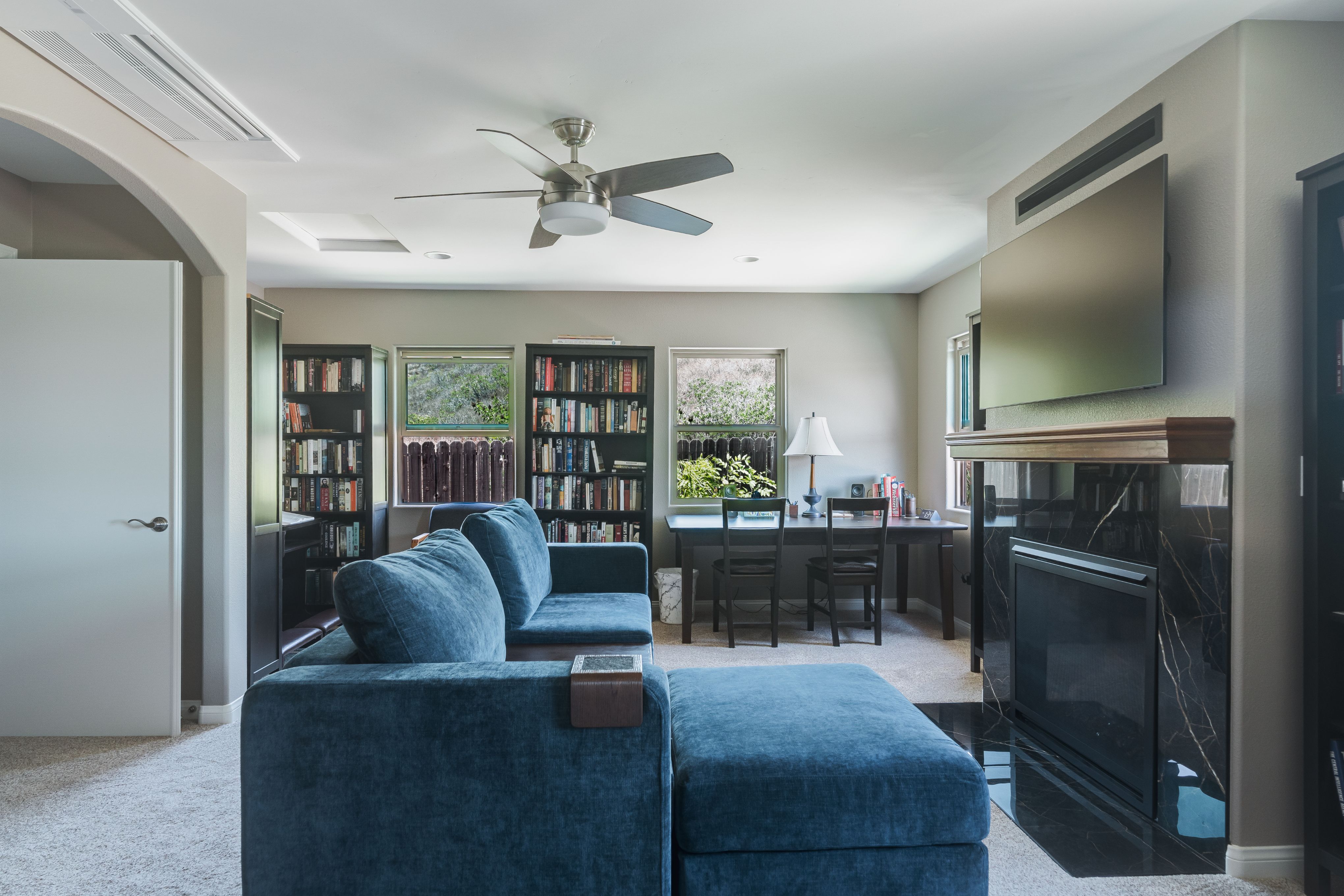 Library and media room with dark wood bookcases, writing desk, wall-mounted TV, fireplace and ceiling fan over carpet