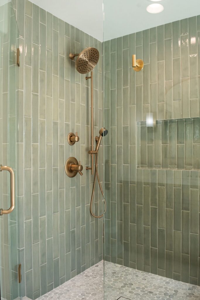 Walk-in shower with green vertical tile, gold-tone rain head, handheld sprayer on slide bar, tiled niche and pebble floor