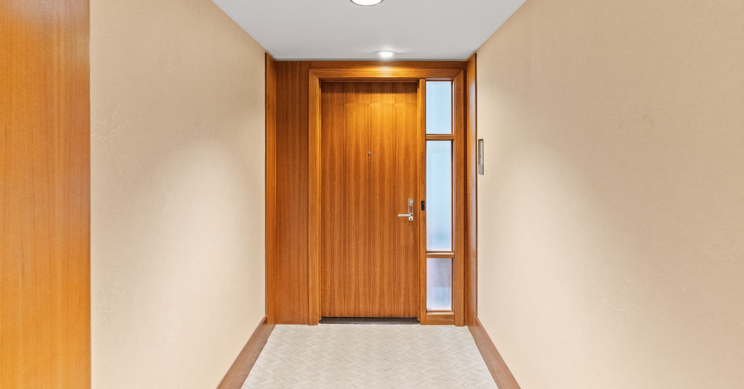 Interior hallway leading to a wood front door with a narrow glass sidelight, recessed lighting, and wood trim.