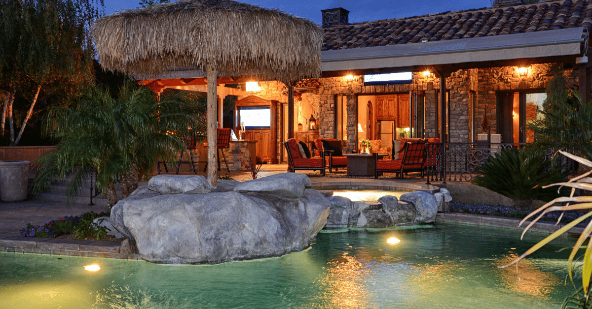 Pool at dusk with a rock waterfall, a thatched palapa, a raised spa and a stone home with a lit covered patio, outdoor bar and fire table.