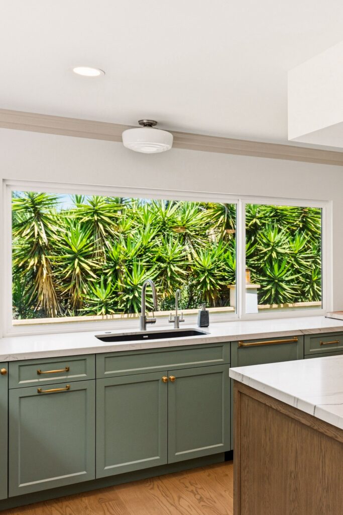 Black undermount sink with stainless faucet and second tap below a wide window, set in green cabinets with brass hardware